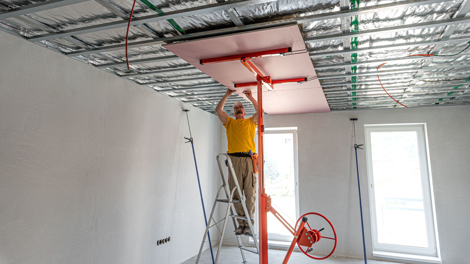 The worker screwing plasterboard to the ceiling. He is using special electric screwdriver and drywall lift. A red plasterboard improve the fire resistance of ceiling structures.